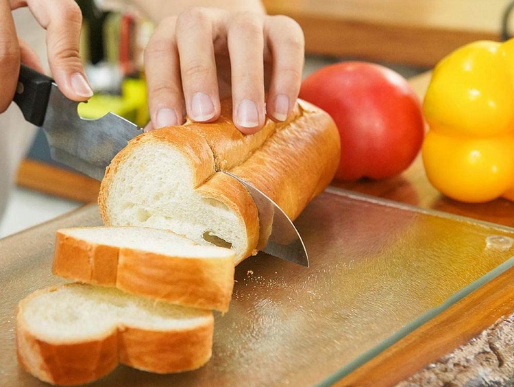 loaf of bread on cutting board with peppers beside it and human hand cutting