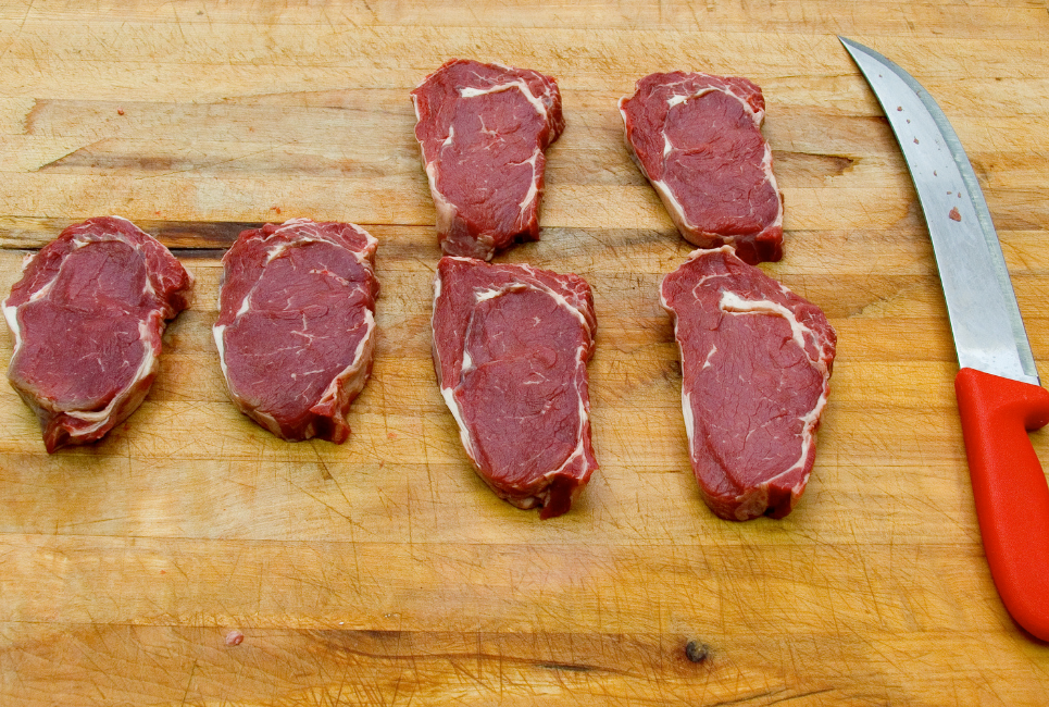 six raw steaks on butcher block cutting board