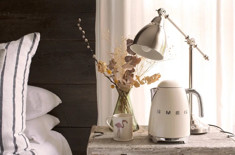 SMEG brand white electric tea kettle on nightstand with lamp flowers and flamingo print mug