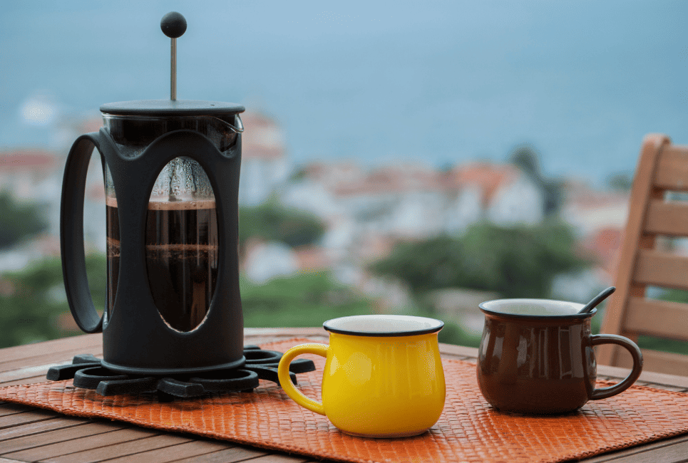black french press coffee maker on table outside next to two cups of coffee
