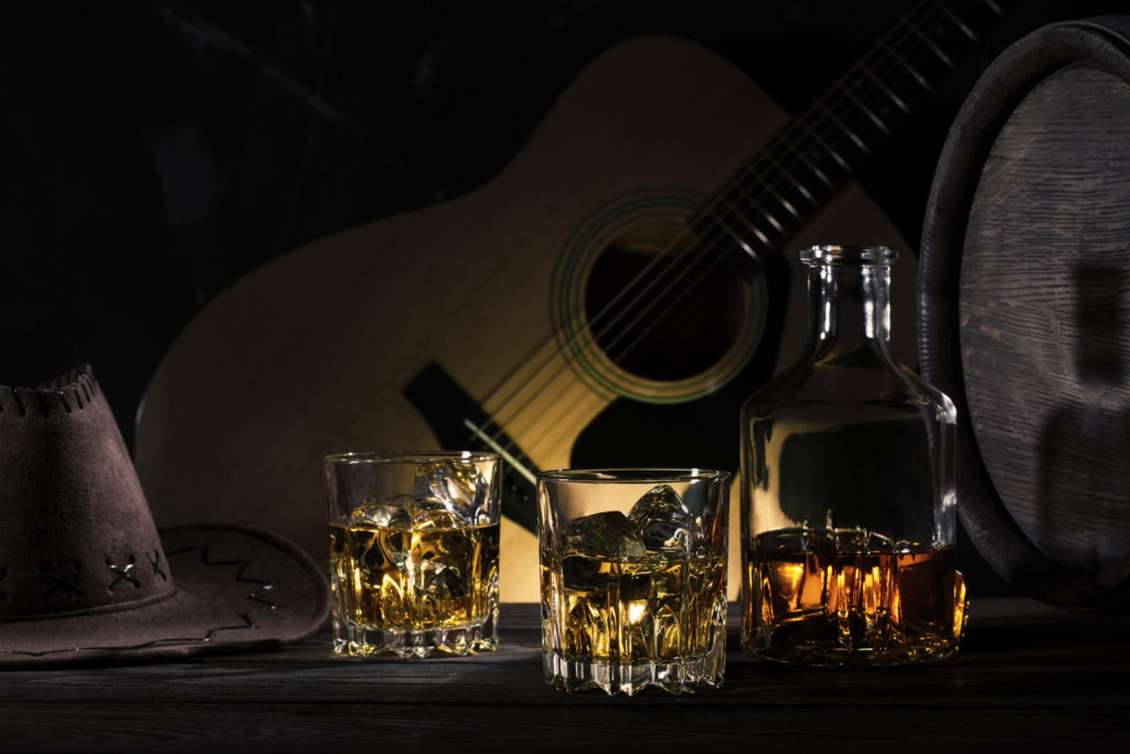 Two glasses of whiskey with ice and cowboy hat on wooden table on guitar background
