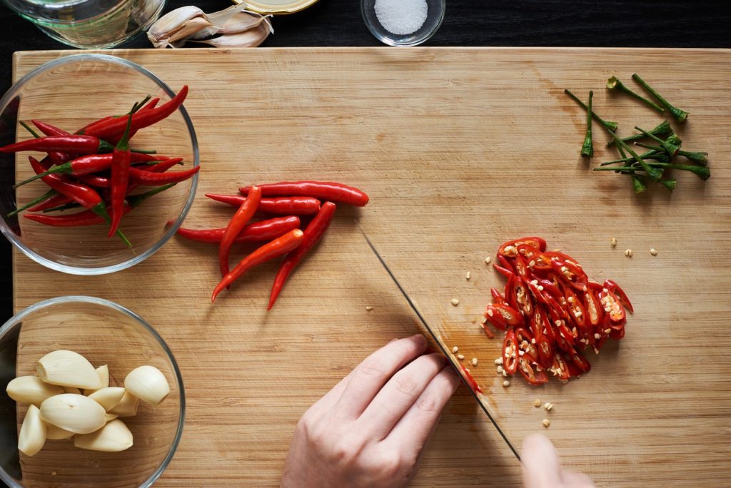 persons hands slicing chili peppers on cutting board with bowls of peppers and garlic around