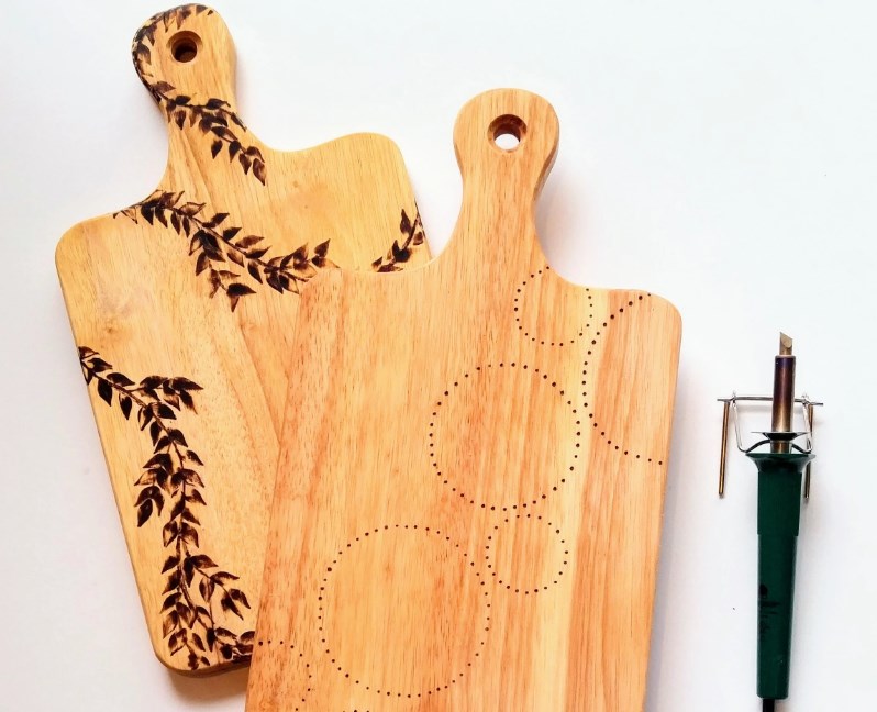two cutting boards with burnt in floral and dainty design and pyrograph tool next to them